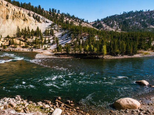 A serene river curves through a forested landscape, surrounded by rocky hills and clear blue skies.