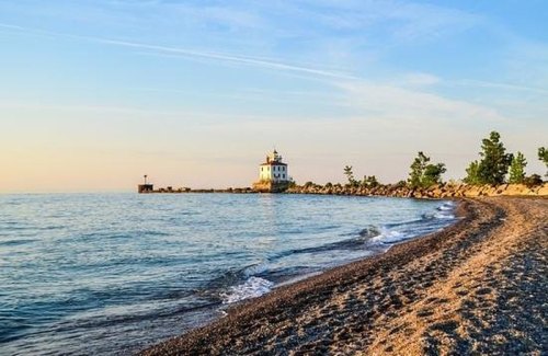 A serene beach scene with gentle waves, a lighthouse in the distance, and a clear sky at sunset.