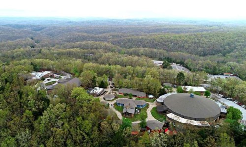 Aerial view of a wooded area with buildings nestled among trees, surrounded by rolling hills and a cloudy sky.