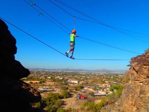 A person in a bright green shirt balances on a tightrope high above a scenic landscape with mountains in the distance.