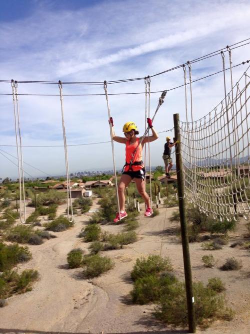 A person in a helmet and harness navigates a rope course in a desert landscape, with another participant in the background.