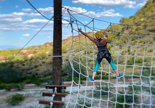 A person in a yellow helmet climbs a rope net against a scenic outdoor backdrop with blue skies and greenery.