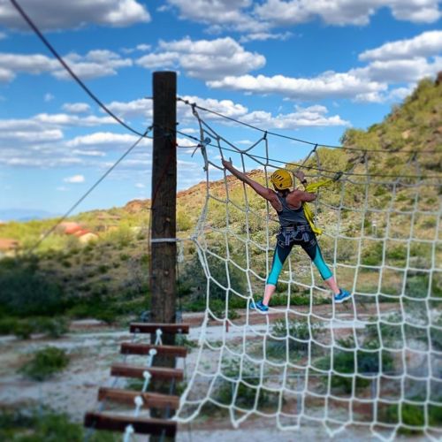 A person climbs a rope net against a backdrop of blue skies and green hills, wearing a helmet and climbing gear.