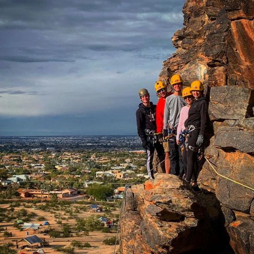 A group of five climbers in helmets stands on a rocky cliff, overlooking a valley and distant cityscape.