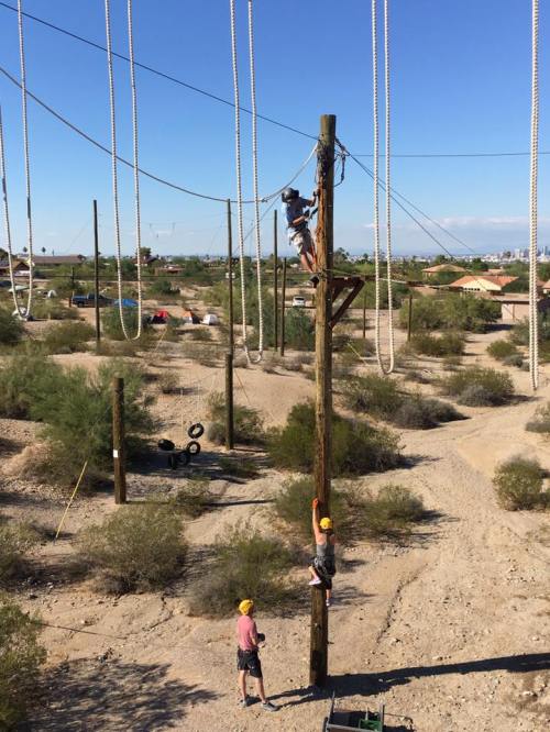 Two people in helmets work on a utility pole in a desert area, with ropes and equipment visible in the background.