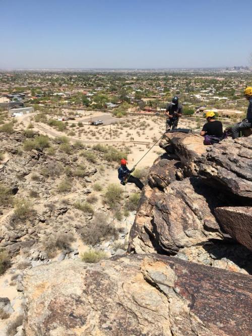 A person rappels down a rocky cliff with a desert landscape and city in the background, while others observe nearby.
