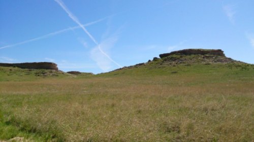 A wide grassy landscape with two rocky outcrops under a clear blue sky with wispy clouds.