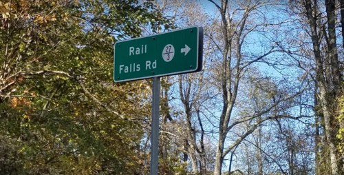 Green road sign indicating "Rail" and "Falls Rd" with a directional arrow, surrounded by trees.