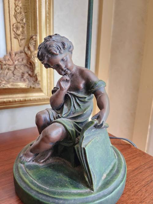 A bronze statue of a seated child reading, resting on a circular base, with a decorative frame in the background.