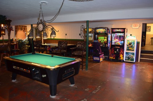 A cozy game room featuring a pool table, arcade machines, and seating areas with dim lighting.