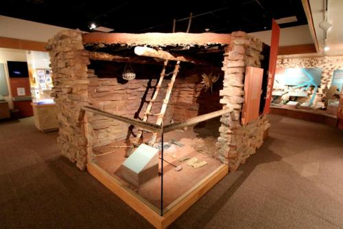 A stone structure resembling a historical dwelling, displayed in a museum with artifacts and informational panels nearby.