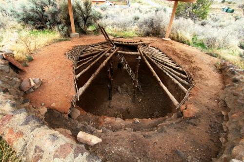 An underground structure with a circular shape, made of wooden beams and surrounded by earthy terrain and sparse vegetation.