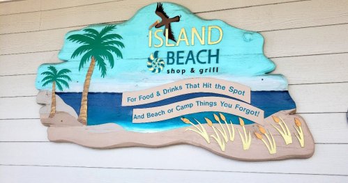 Colorful sign for "Island Beach Shop & Grill" featuring palm trees, ocean, and text about food and beach essentials.