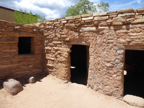 A weathered adobe structure with stone walls and doorways, set in a sandy area under a partly cloudy sky.