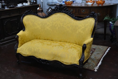 A vintage yellow upholstered sofa with ornate black wooden frame, set in an antique shop.