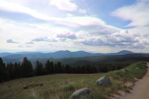 A scenic view of rolling mountains and a cloudy sky, with a dirt path and trees in the foreground.
