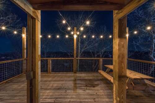 A wooden deck at night, illuminated by string lights, with a view of trees and distant city lights.