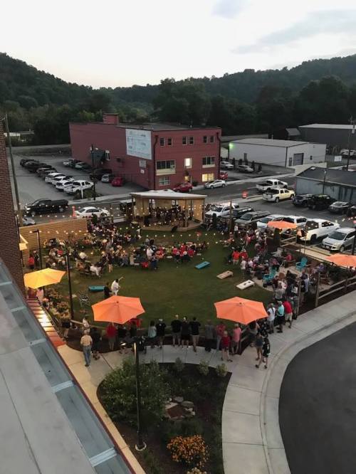 Aerial view of a lively outdoor gathering with people seated under orange umbrellas in a green space at dusk.