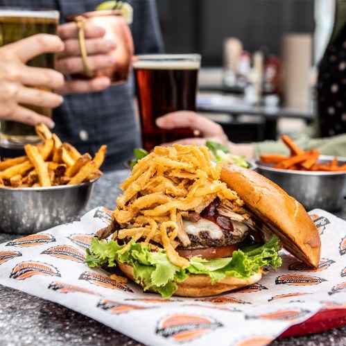 A close-up of a burger topped with crispy onions, lettuce, and cheese, with fries and drinks in the background.