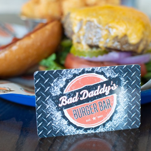 A close-up of a "Bad Daddy's Burger Bar" gift card in front of a burger and bun on a blue plate.