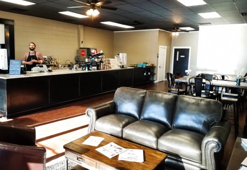 A cozy café interior with a barista at the counter, seating area, and wooden tables. Bright and inviting atmosphere.