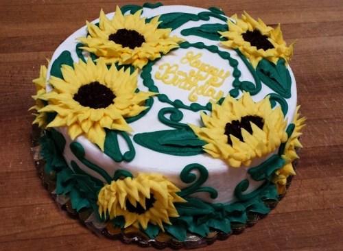 A round birthday cake decorated with yellow sunflowers and green leaves, with "Happy Birthday" written on top.