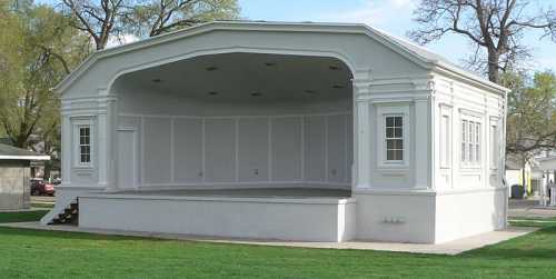 A white, open-air bandstand with a curved roof and steps, set in a grassy park area.