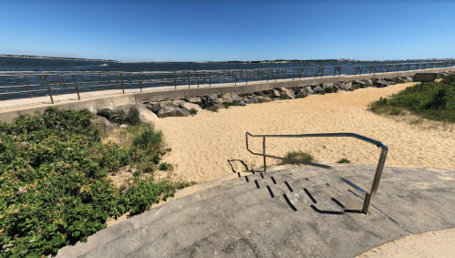 A sandy beach area with a pathway and railing along the water, surrounded by greenery and a clear blue sky.