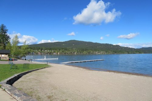 A serene lakeside view with a sandy beach, dock, and green hills under a blue sky with fluffy clouds.