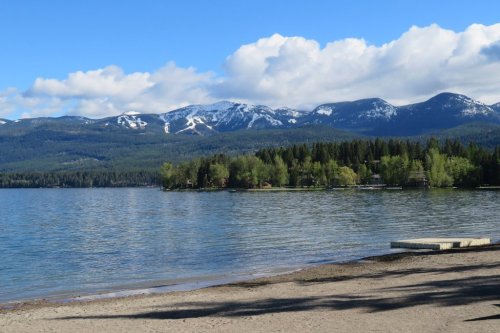 A serene lake surrounded by lush trees and snow-capped mountains under a blue sky with fluffy clouds.