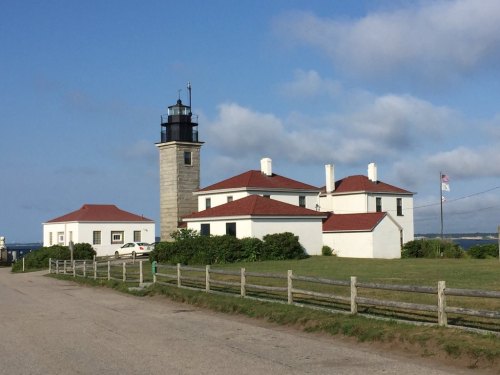 A white lighthouse with a black lantern, surrounded by buildings and greenery, under a blue sky with scattered clouds.