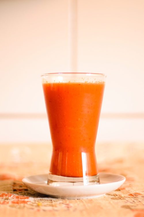A glass of vibrant orange juice sits on a small white plate against a light background.