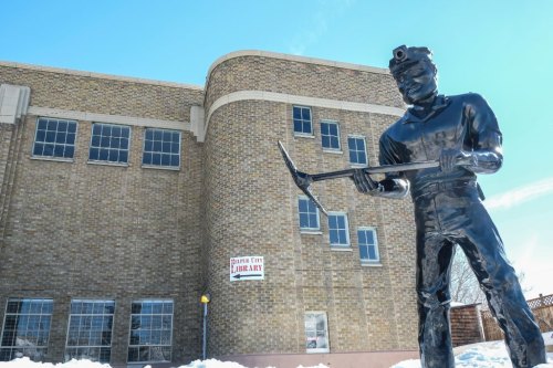 A black metal statue of a miner holding a pickaxe stands in front of a brick building with a library sign.