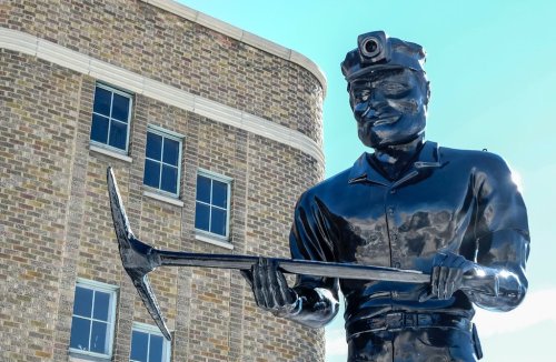 A black statue of a miner holding a pickaxe, with a building featuring large windows in the background.