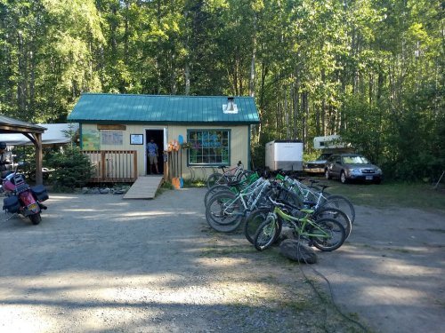 A small building with a green roof surrounded by trees, featuring parked bicycles and cars in a gravel area.