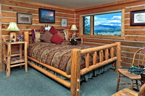 Cozy log cabin bedroom with a wooden bed, decorative pillows, lamps, and a scenic window view of mountains.