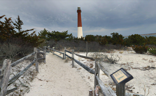 A sandy path leads to a lighthouse, surrounded by greenery and under a cloudy sky. A sign is visible along the path.