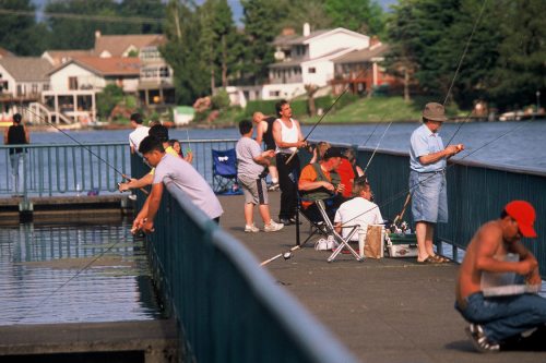 People fishing from a pier by a lake, with houses in the background and a clear blue sky.