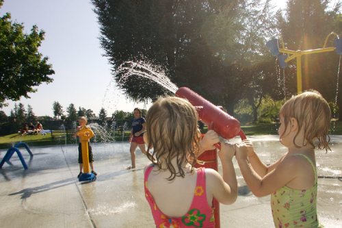 Children play in a splash pad, enjoying water sprays on a sunny day, surrounded by trees and grassy areas.