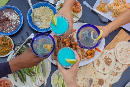 A group toasts with colorful drinks over a spread of food, including grilled meats, tortillas, and various sides.