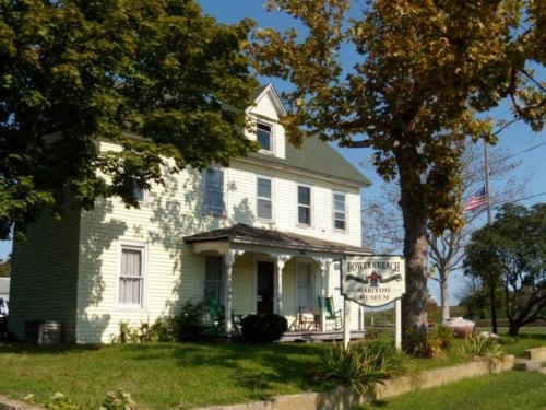 A yellow, two-story house with a porch, labeled as Bowser Beach Maritime Museum, surrounded by trees and blue sky.