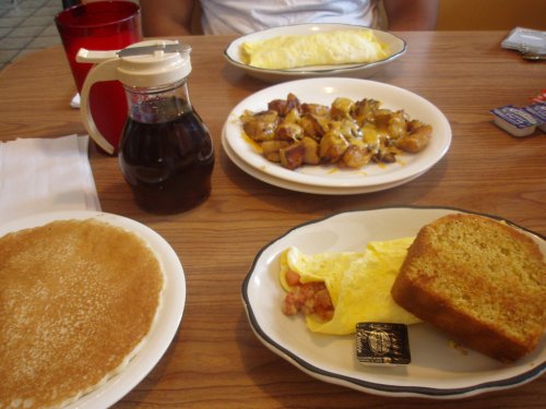 A breakfast spread featuring pancakes, an omelet, cheesy potatoes, cornbread, and syrup on a table.