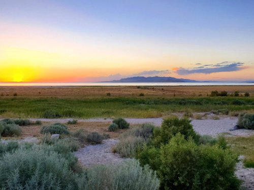 Sunset over a serene landscape with grassy fields and distant mountains reflecting on calm waters.