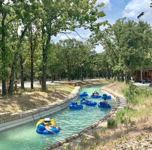 People float on blue inner tubes in a winding waterway surrounded by trees and a clear sky.