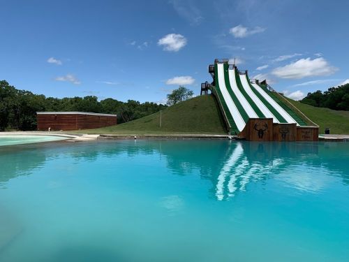 A large water slide on a grassy hill beside a clear blue pool under a bright sky with a few clouds.
