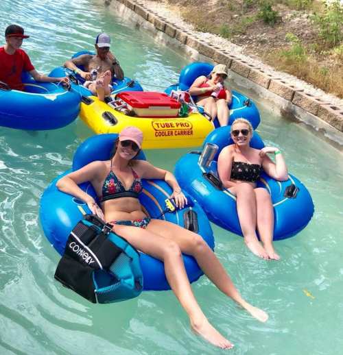 Two women in swimsuits relax on blue inner tubes in a lazy river, enjoying the sun and water. Others float nearby.