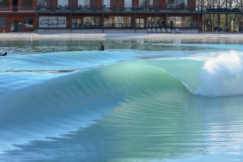 A smooth, turquoise wave rolls in at a surf park, with a surfer riding the crest in the background.