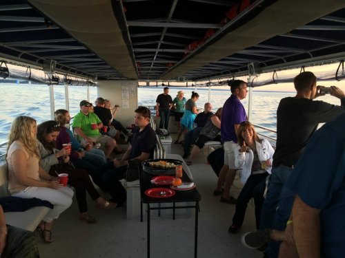 A group of people socializing on a boat, enjoying drinks and snacks while overlooking the water at sunset.