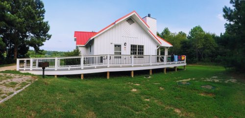 A white house with a red roof, surrounded by greenery, featuring a large deck and a scenic view.