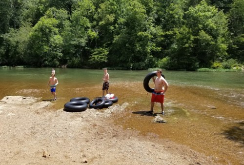 Three boys stand by a river, holding inner tubes, with lush greenery in the background on a sunny day.